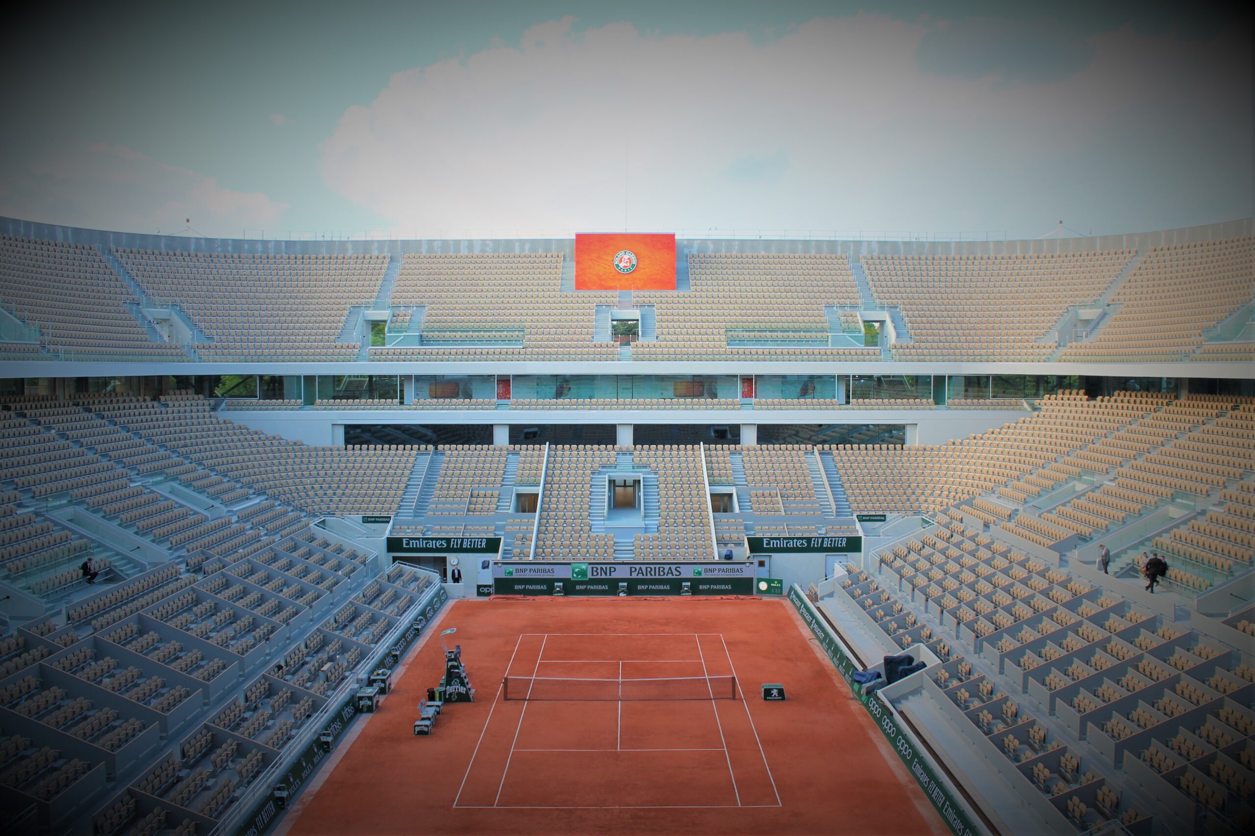 Terrain de tennis rouge au centre du stade, tribunes vides et drapeau orange en hauteur, ambiance calme et lumineuse. super tie-break roland-garros évoqué.