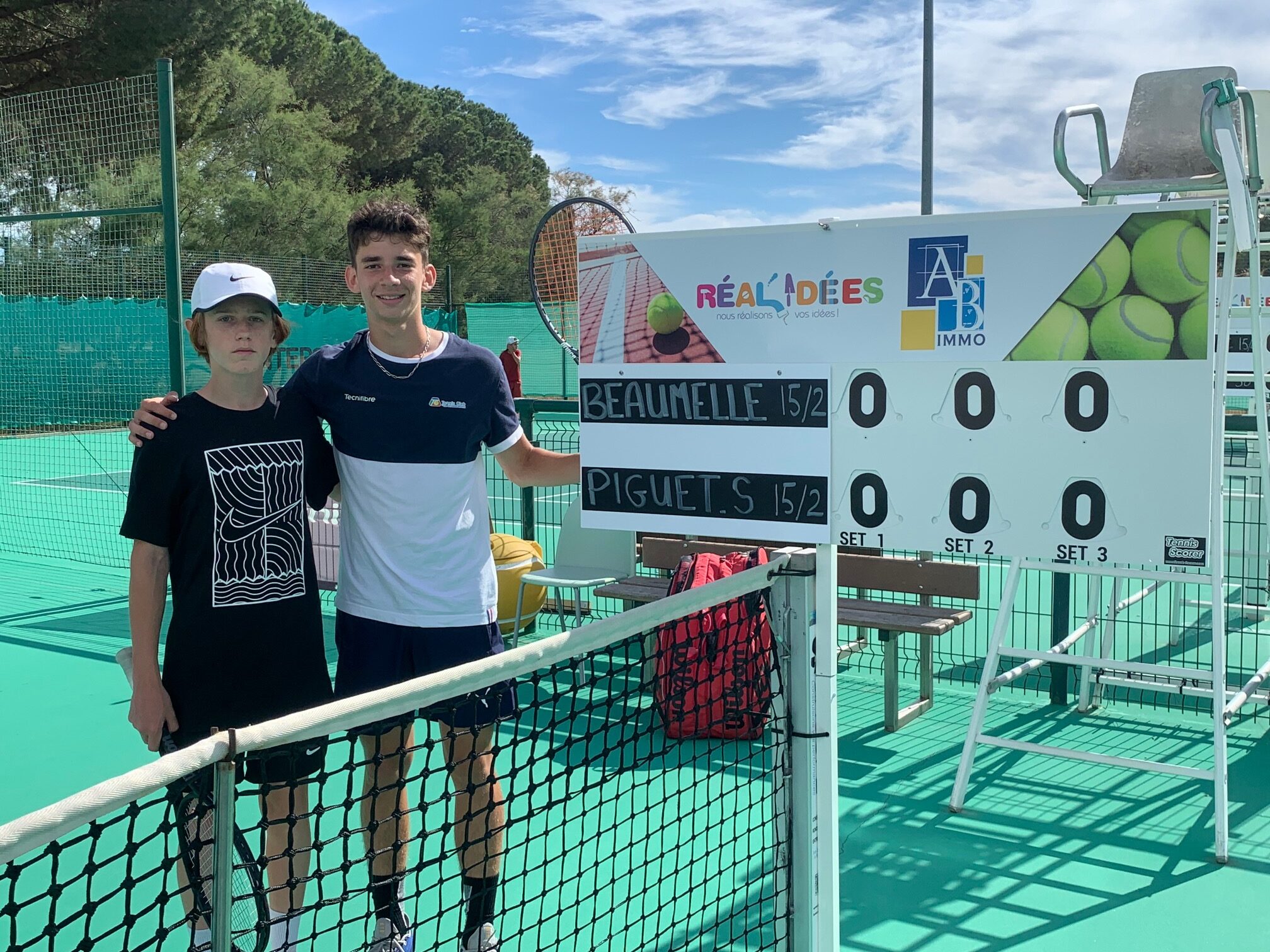 Zwei junge Tennisspieler stehen neben einem Spielstands-Board auf dem Tennisplatz, lachend, mit Netz im Vordergrund.