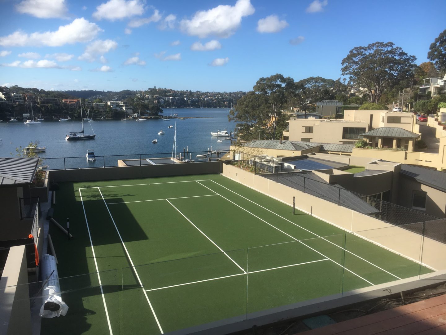 Blick auf eine grünspielfeld Tennis- oder Tennisplatzfläche mit Glaszaun und umliegenden Gebäuden am Wasser.