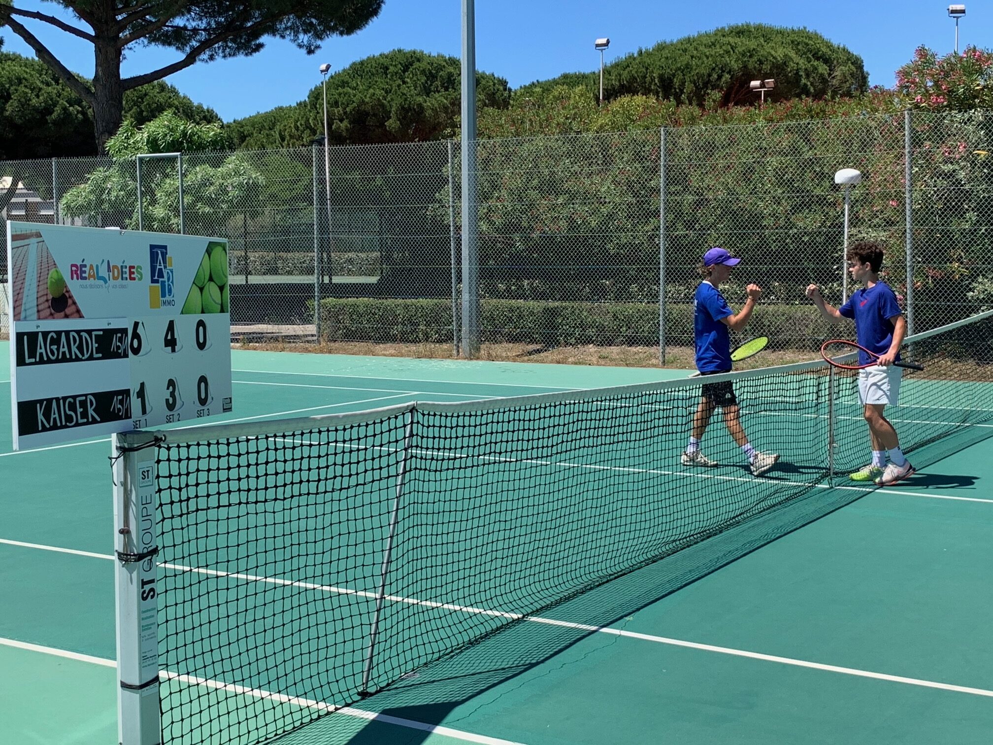 Dos jugadores de tenis conversan junto a la red en una pista al aire libre, con un marcador de puntuación a la izquierda.