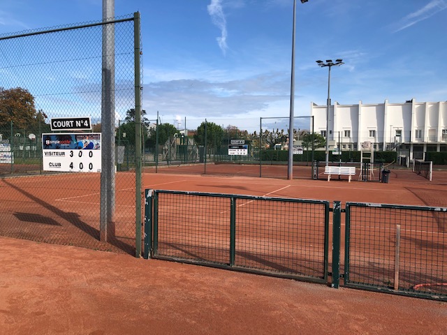 Outdoor tennis court complex with a clay court, fencing, and a scoreboard visible.
