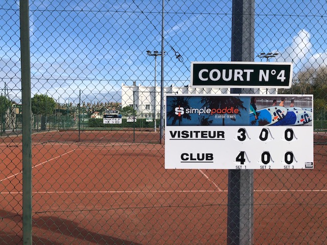 A tennis court with a scoreboard mounted on a fence showing Court No4 and the scores Visitors 3, Club 4.