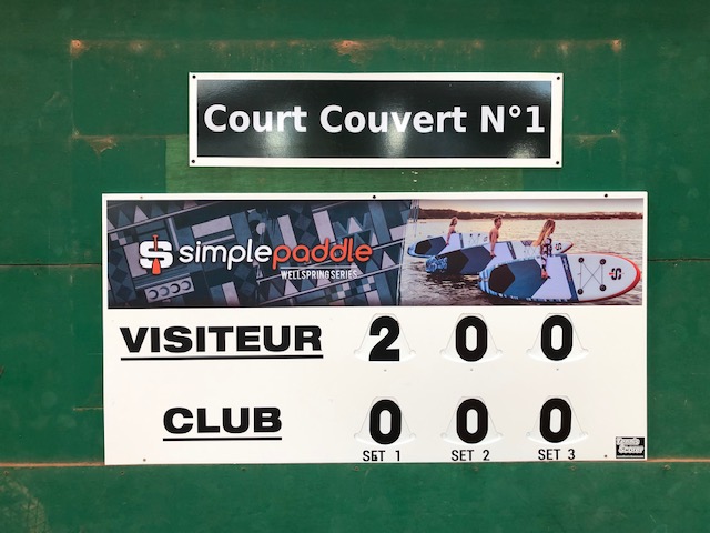 Scoreboard showing visitors 2-0 against club, mounted on a green wall.
