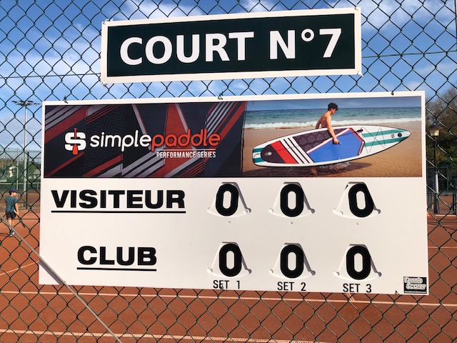 Scoreboard on a tennis court fence showing visitors and club names with zeroes.