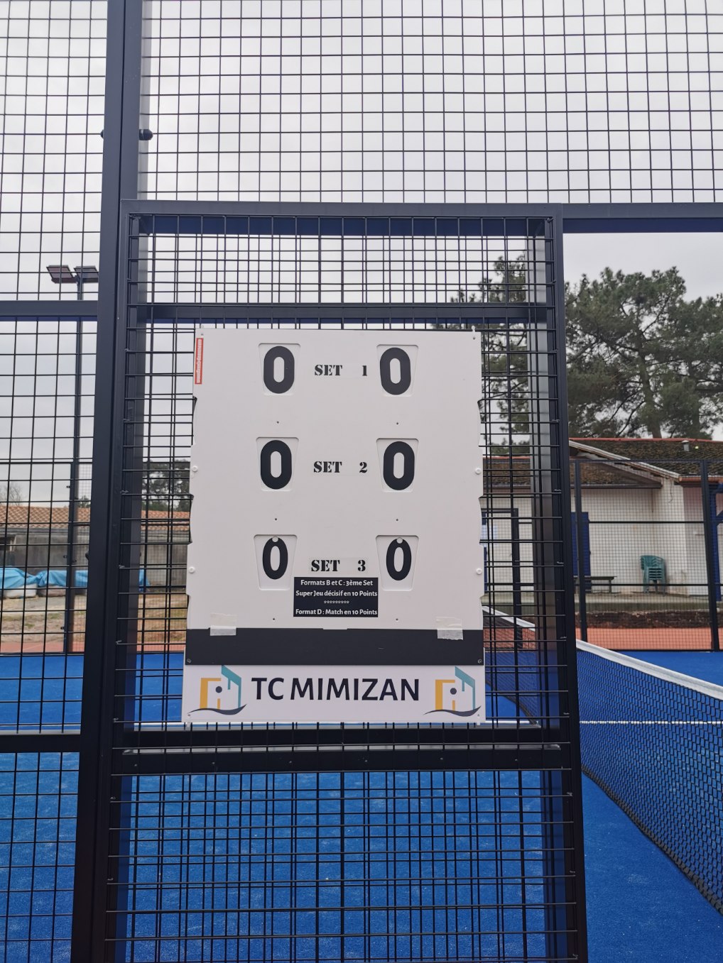 Tableau d’un score de tennis affichant 0–0 pour chaque set sur un terrain extérieur entouré de grillage.