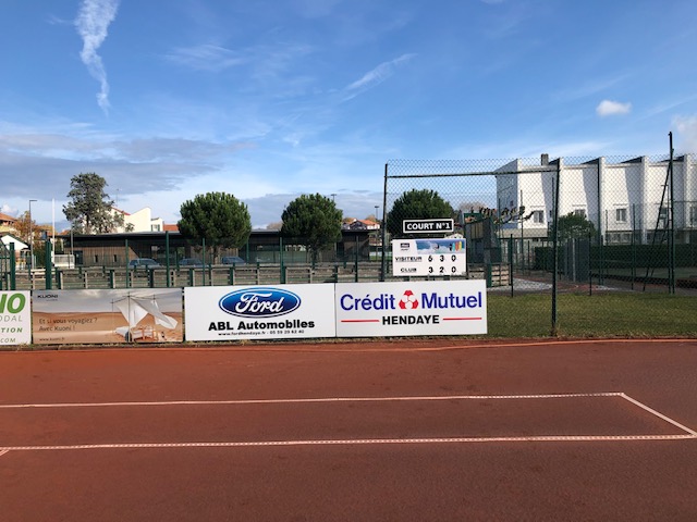 Tennis court with advertising boards along the side and a scoreboard in the background under a blue sky.