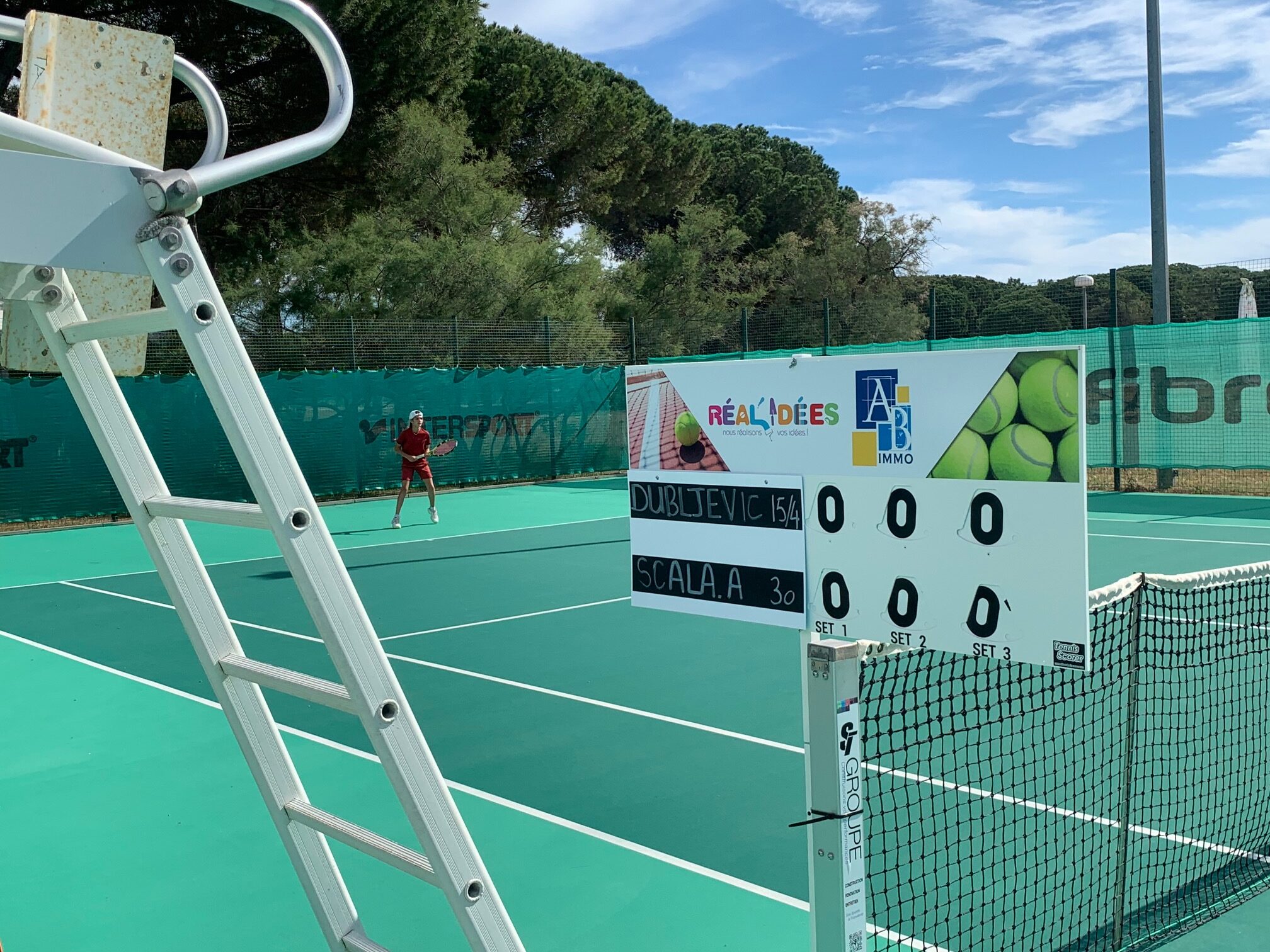 A tennis court with a scoreboard and umpire chair in the foreground, players on the court and a fence with trees in the